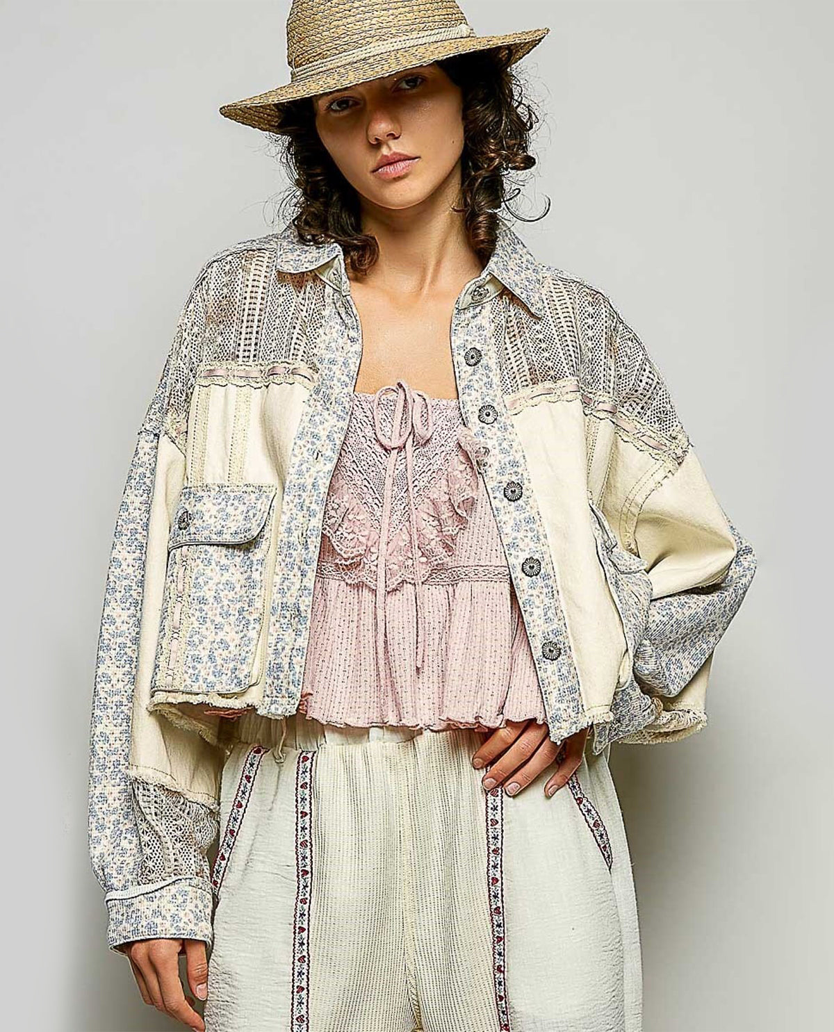 A model wearing a bohemian-style patchwork jacket with cream and blue floral prints, a pink ruffled top, matching cream trousers, and a straw hat, photographed against a neutral gray background.