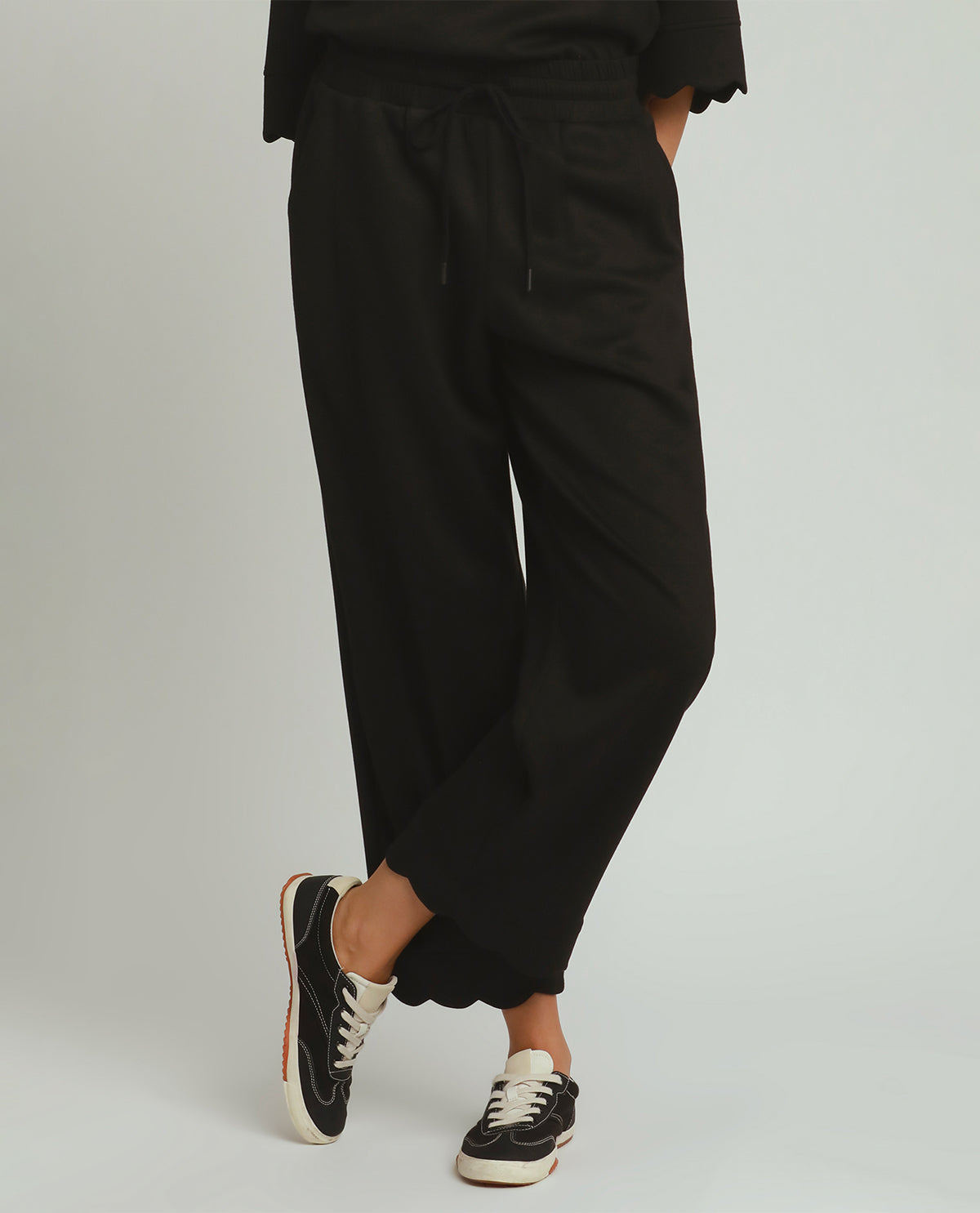 A person wearing loose black drawstring pants with a relaxed, wide-leg fit, paired with black sneakers, shown from the waist down against a light gray studio background.