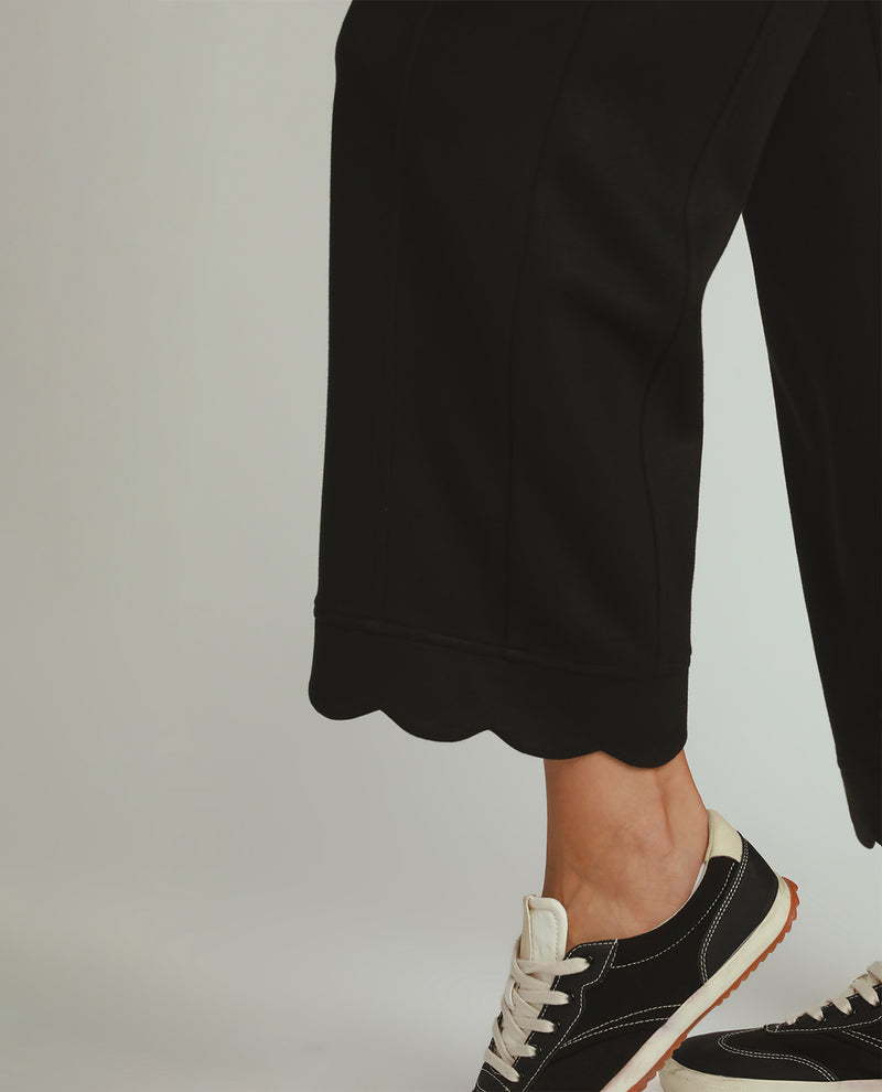 Close-up of a person's lower legs wearing black cropped pants with a scalloped hem and black low-top sneakers against a light gray studio background.