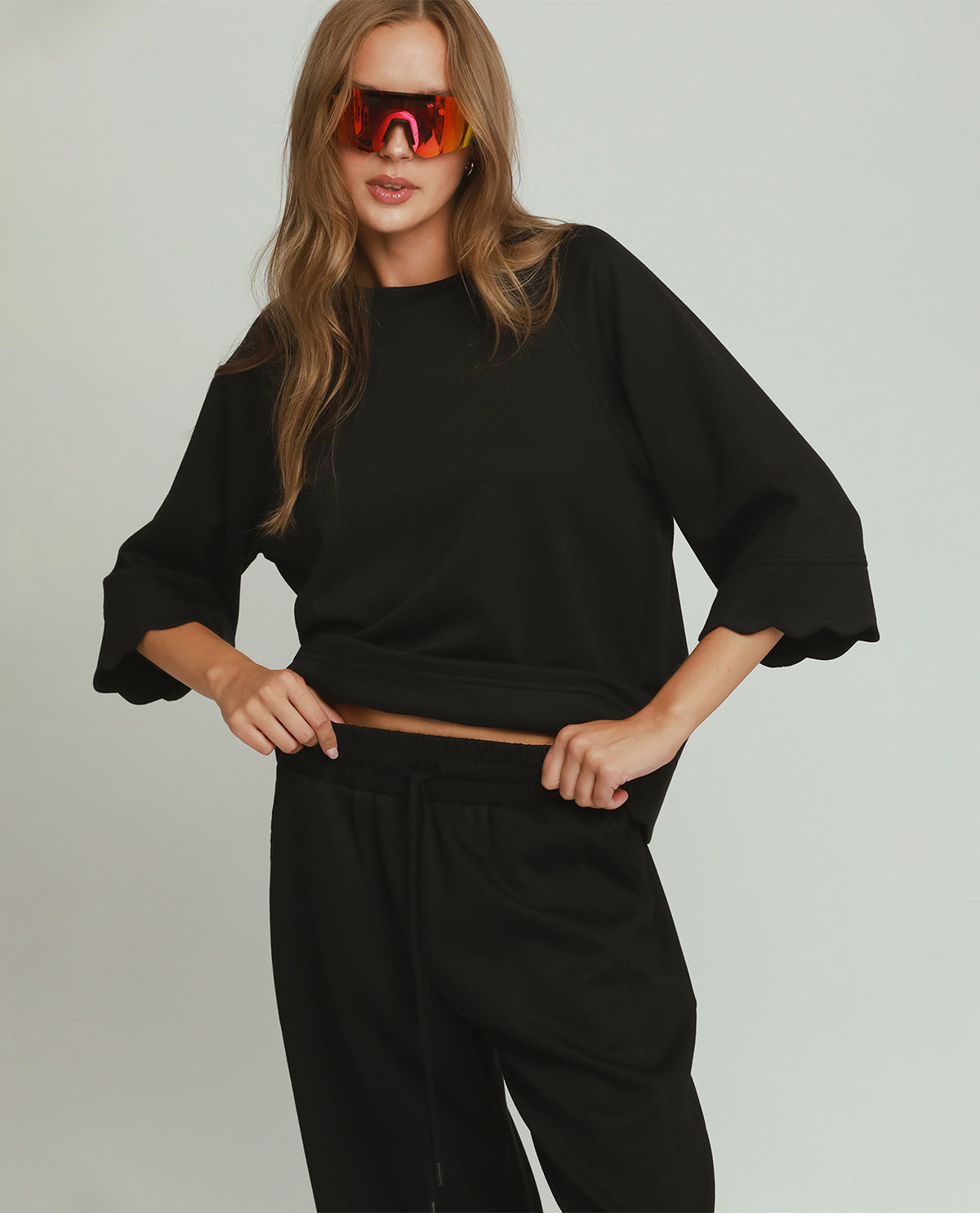 A woman modeling a black oversized crewneck sweatshirt with wide, ruffled three-quarter sleeves and black sweatpants against a light gray studio background.