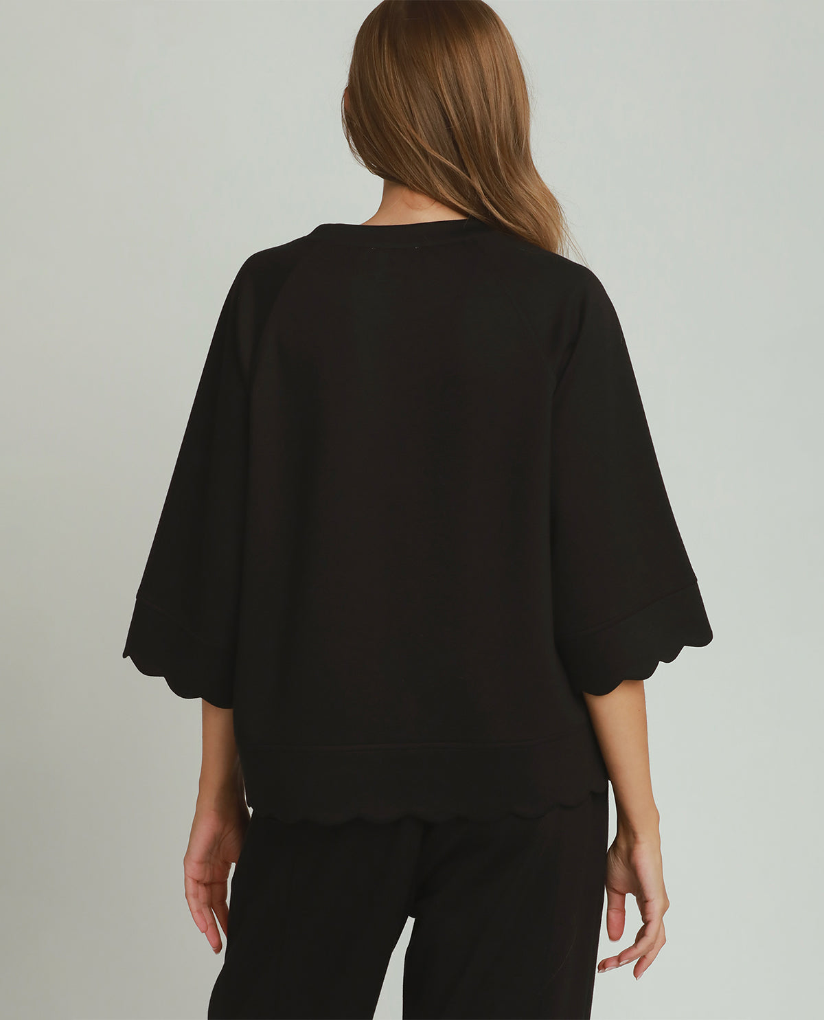A woman shown from the back wearing a black short-sleeve top with scalloped edges and black pants, photographed against a plain light gray studio background.