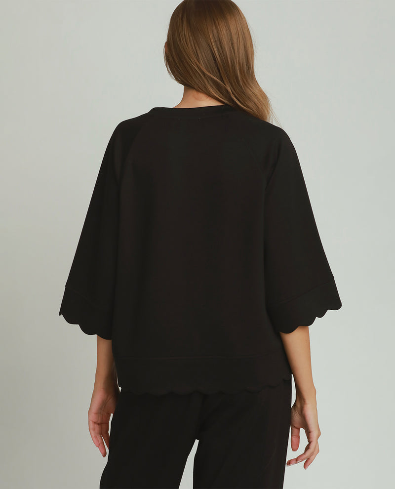 A woman shown from the back wearing a black short-sleeve top with scalloped edges and black pants, photographed against a plain light gray studio background.
