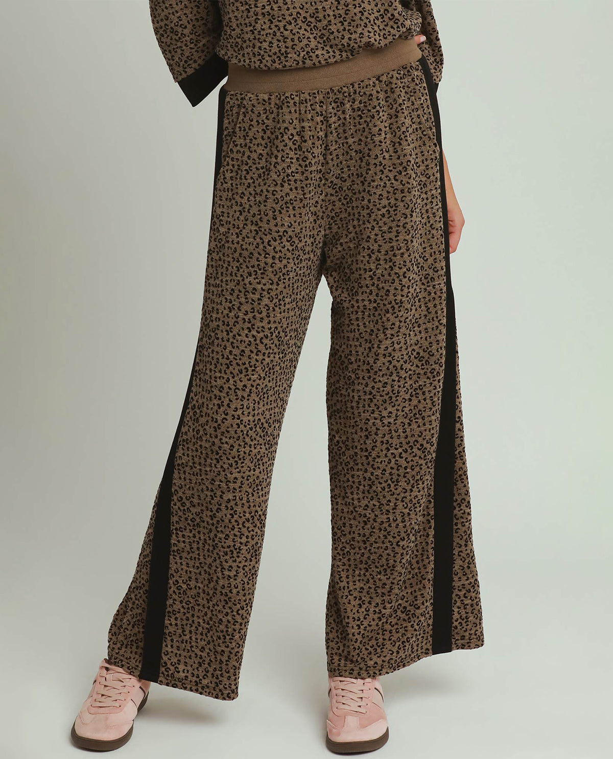 A person wearing brown and black leopard-print wide-leg pants with black side panels, paired with pink sneakers, shown from the waist down against a light gray studio background.