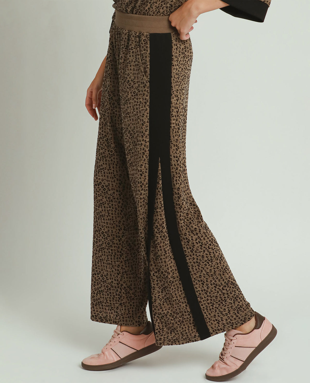 A person wearing wide-leg brown leopard-print pants with a vertical black side stripe and a matching brown belt, paired with light pink sneakers, against a light gray studio background.