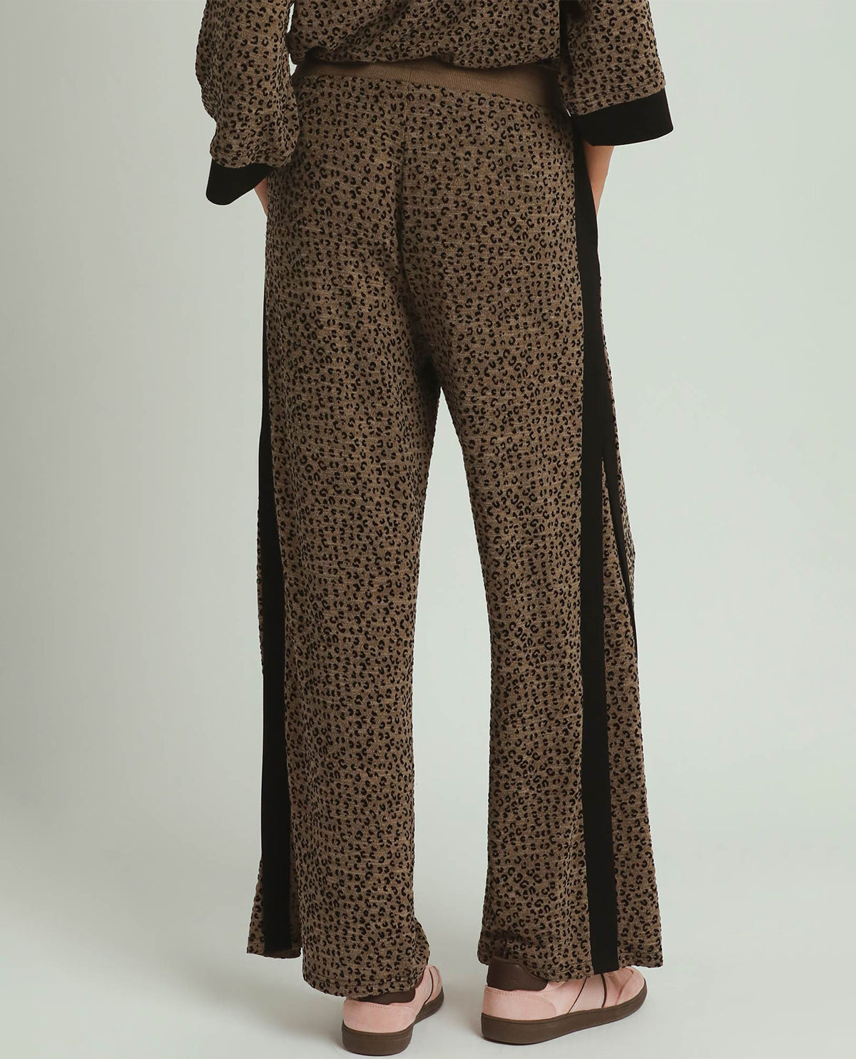 A person shown from the back wearing wide-leg brown pants with a small leopard-like print and black side panels, paired with a matching top and pink sandals, against a light gray studio background.