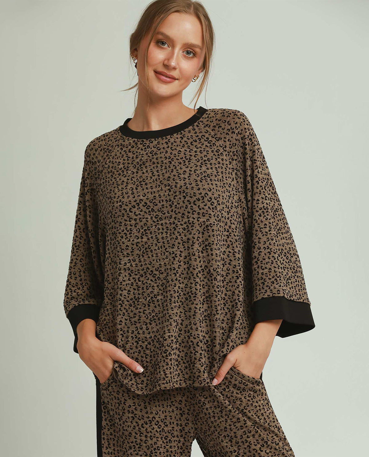 A woman modeling a matching two-piece lounge set featuring a brown and black leopard print pattern with solid black trim on the crew neck, cuffs, and side pockets, set against a solid light gray background.