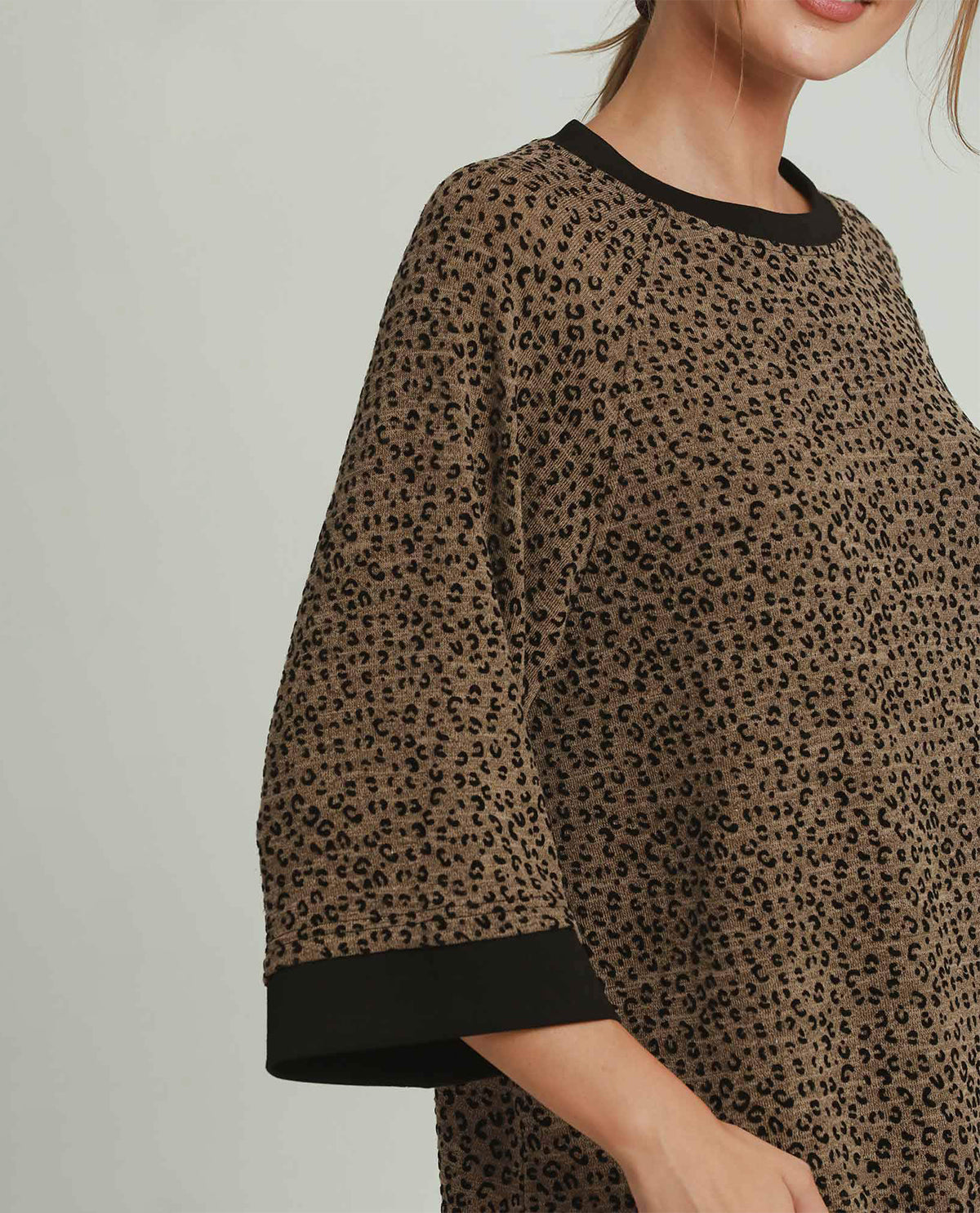 A close-up of a model wearing a brown and black leopard-print tunic top with contrasting black ribbed trim at the crew neck and sleeve cuffs, set against a plain light gray background.
