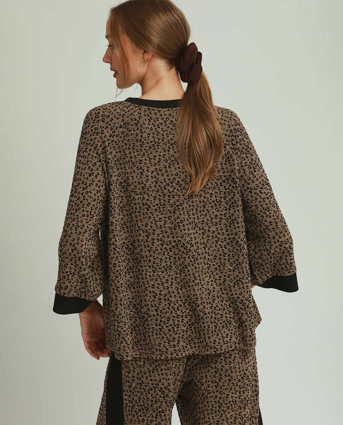 A woman shown from the back wearing a brown and black leopard-print pajama set with solid black trim on the collar, cuffs, and side panels, against a light gray studio background.