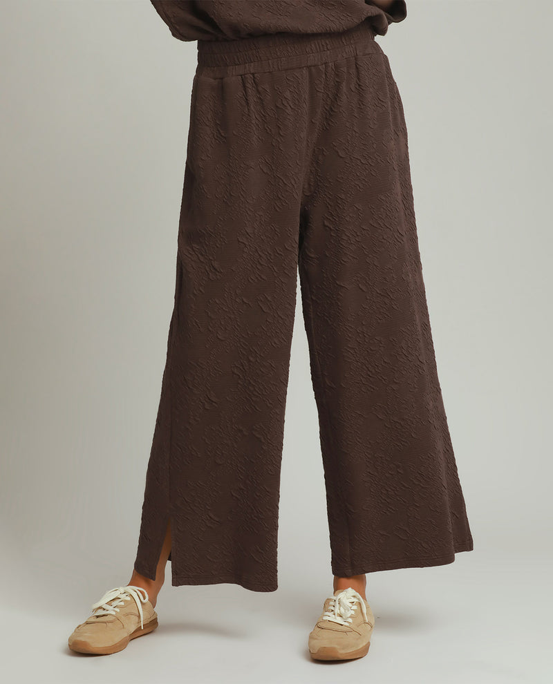 A person wearing dark brown wide-leg knit pants with a textured pattern, shown from the waist down against a light gray studio background. The model is also wearing beige sneakers.