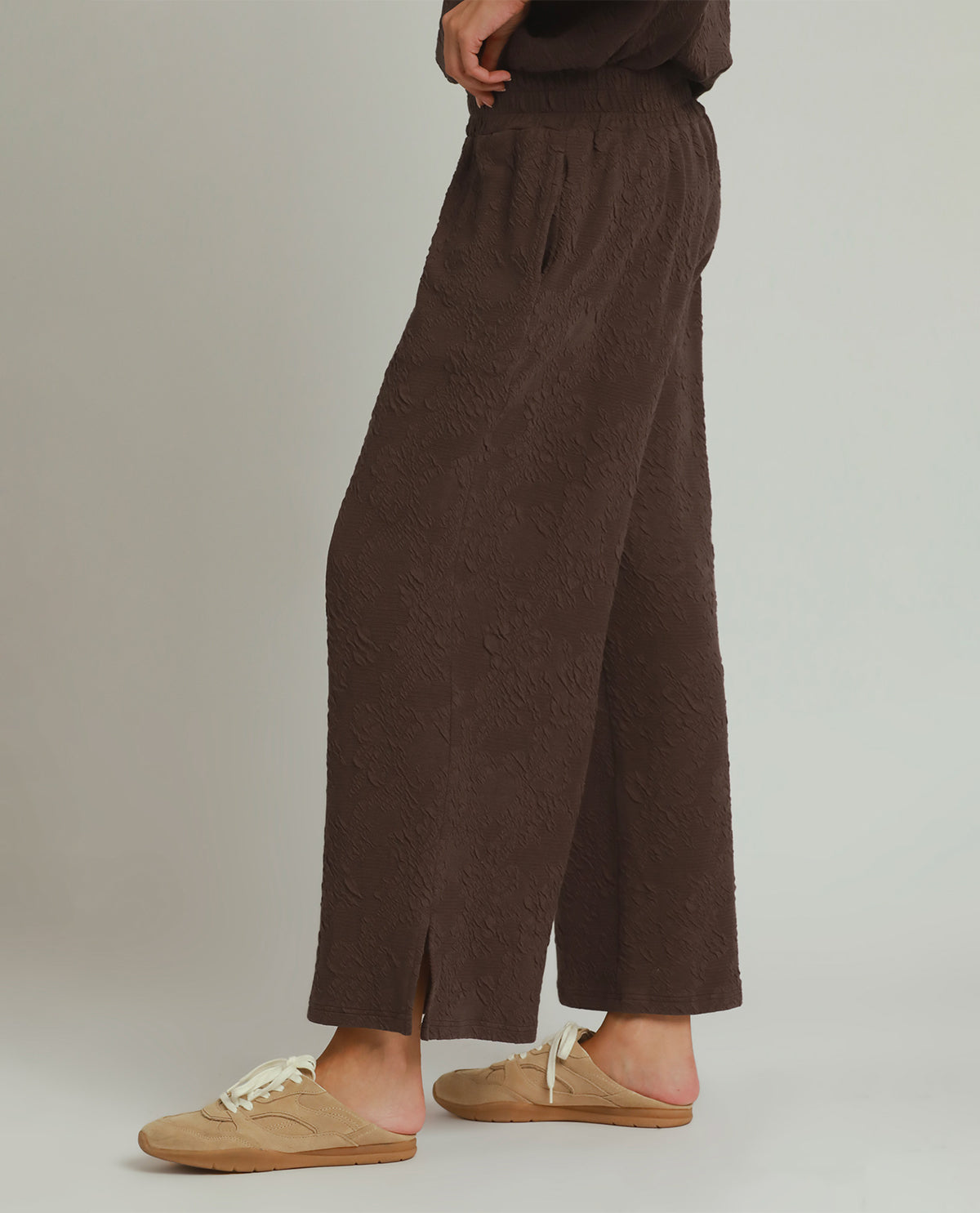 A person wearing dark brown textured wide-leg pants and beige slip-on shoes, shown from the waist down against a plain light gray studio background.
