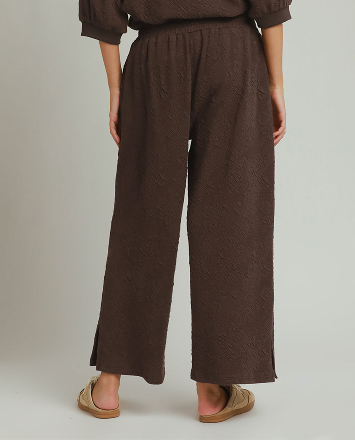 A person shown from the back wearing dark brown textured wide-leg pants and tan slide sandals against a plain light gray studio background.
