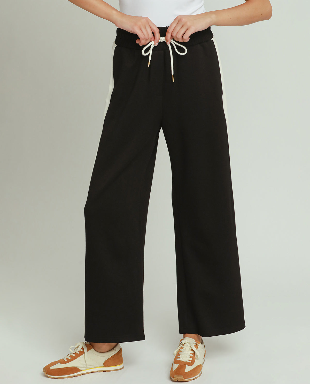 A person wearing black wide-leg drawstring pants with white laces, paired with a white top and tan-and-cream sneakers, shown against a light gray studio background.