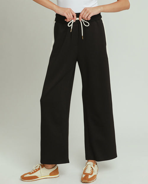 A person wearing black wide-leg drawstring pants with white laces, paired with a white top and tan-and-cream sneakers, shown against a light gray studio background.