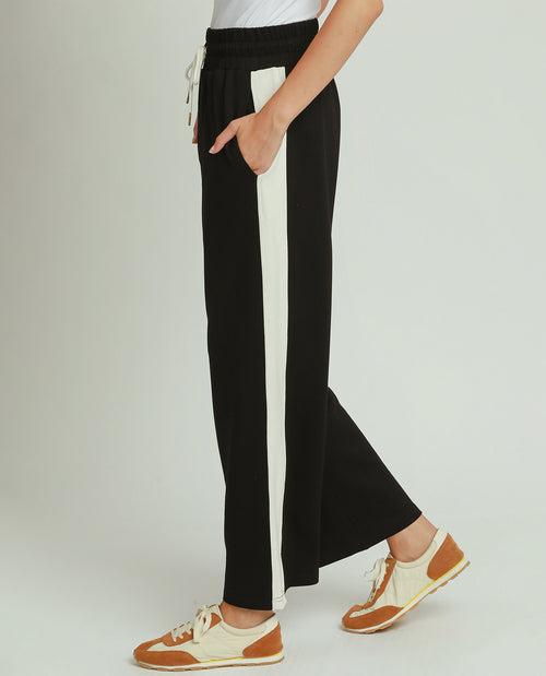 A person wearing black wide-leg pants with a vertical white stripe down the side, paired with brown and cream sneakers, shown against a light gray studio background.
