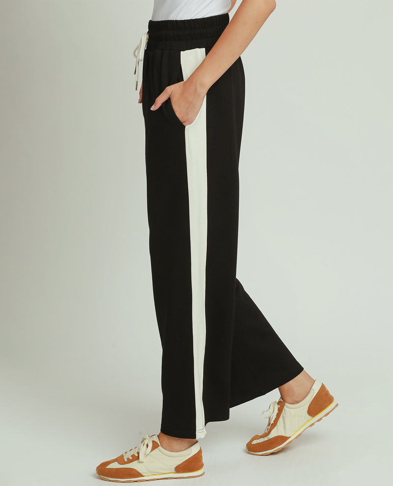 A person wearing black wide-leg pants with a vertical white stripe down the side, paired with brown and cream sneakers, shown against a light gray studio background.