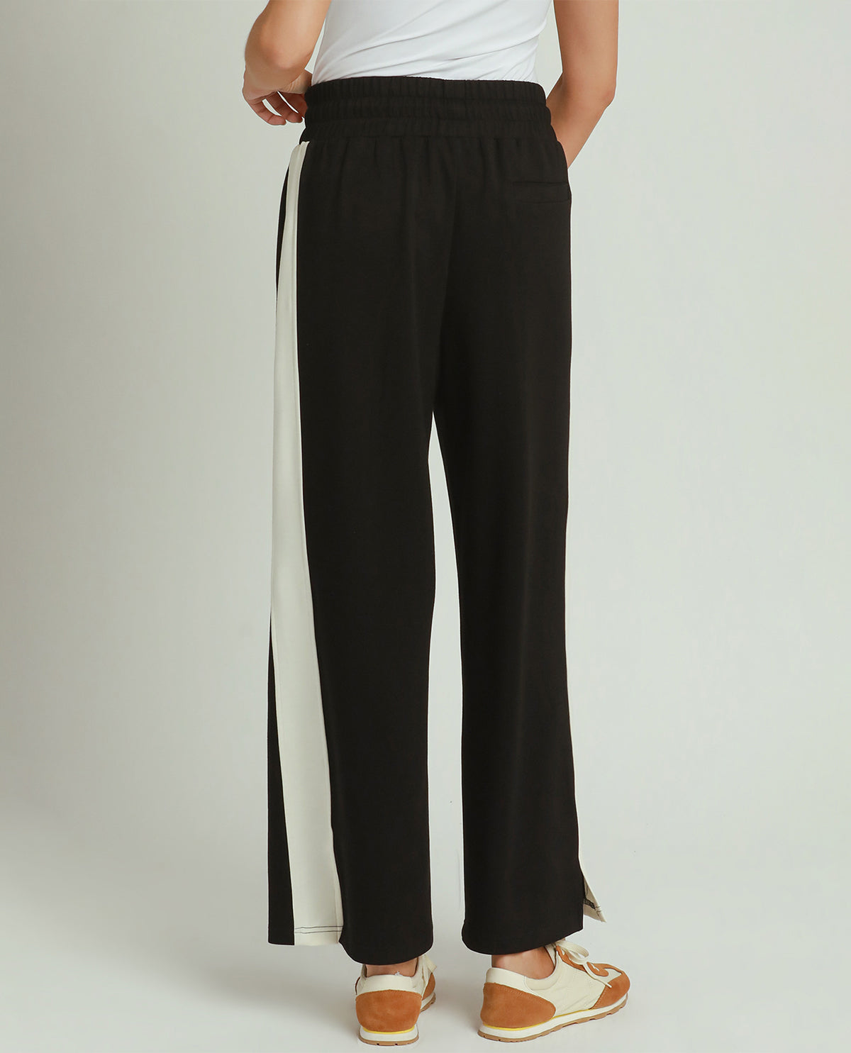 A person shown from the back wearing black wide-leg pants with a wide off-white vertical side stripe, paired with a white top and tan-and-white slip-on shoes, against a light gray studio background.