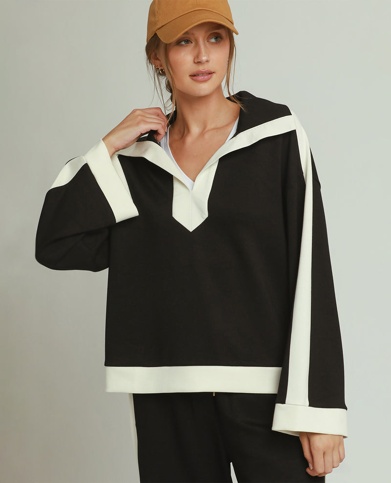 A woman modeling a black long-sleeve top with contrasting white trim details, paired with black pants and a tan baseball cap, against a neutral gray studio background.