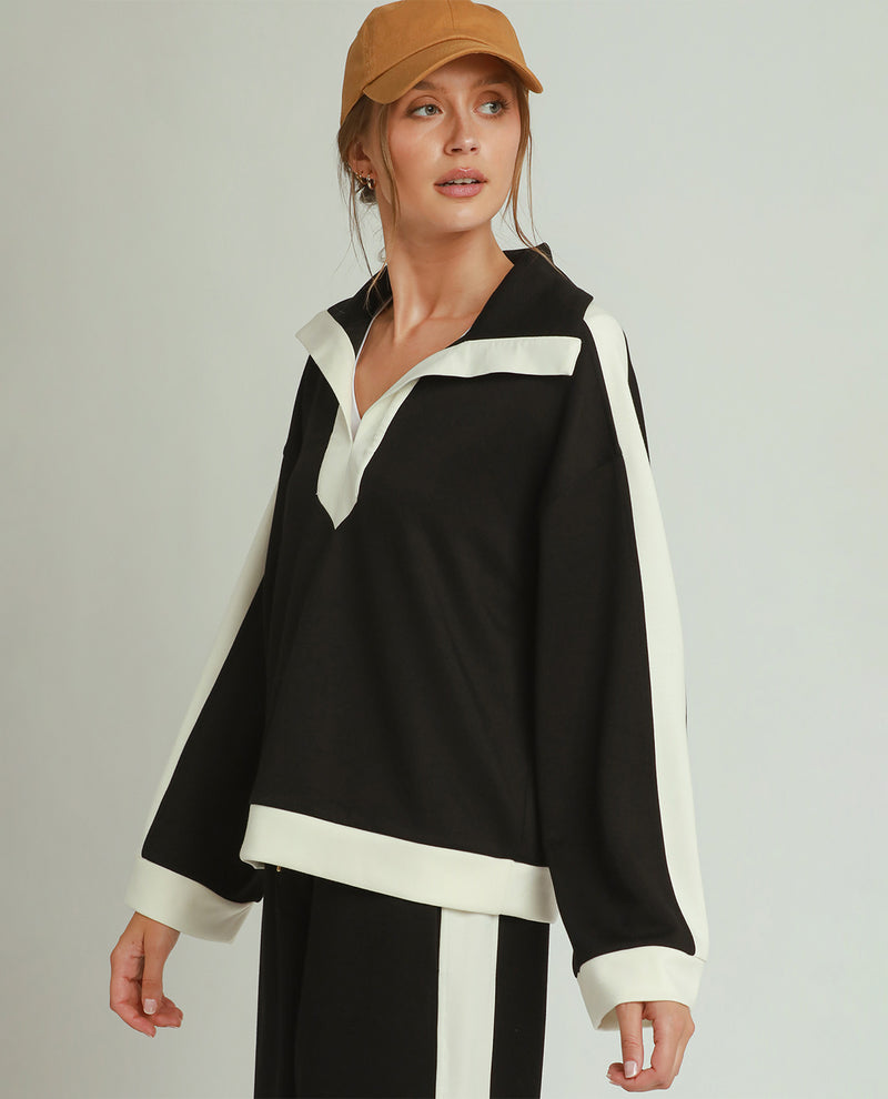 A woman wearing a black long-sleeve half-placket top with contrasting white trim and matching black-and-white pants, paired with a tan cap, against a light gray studio background.
