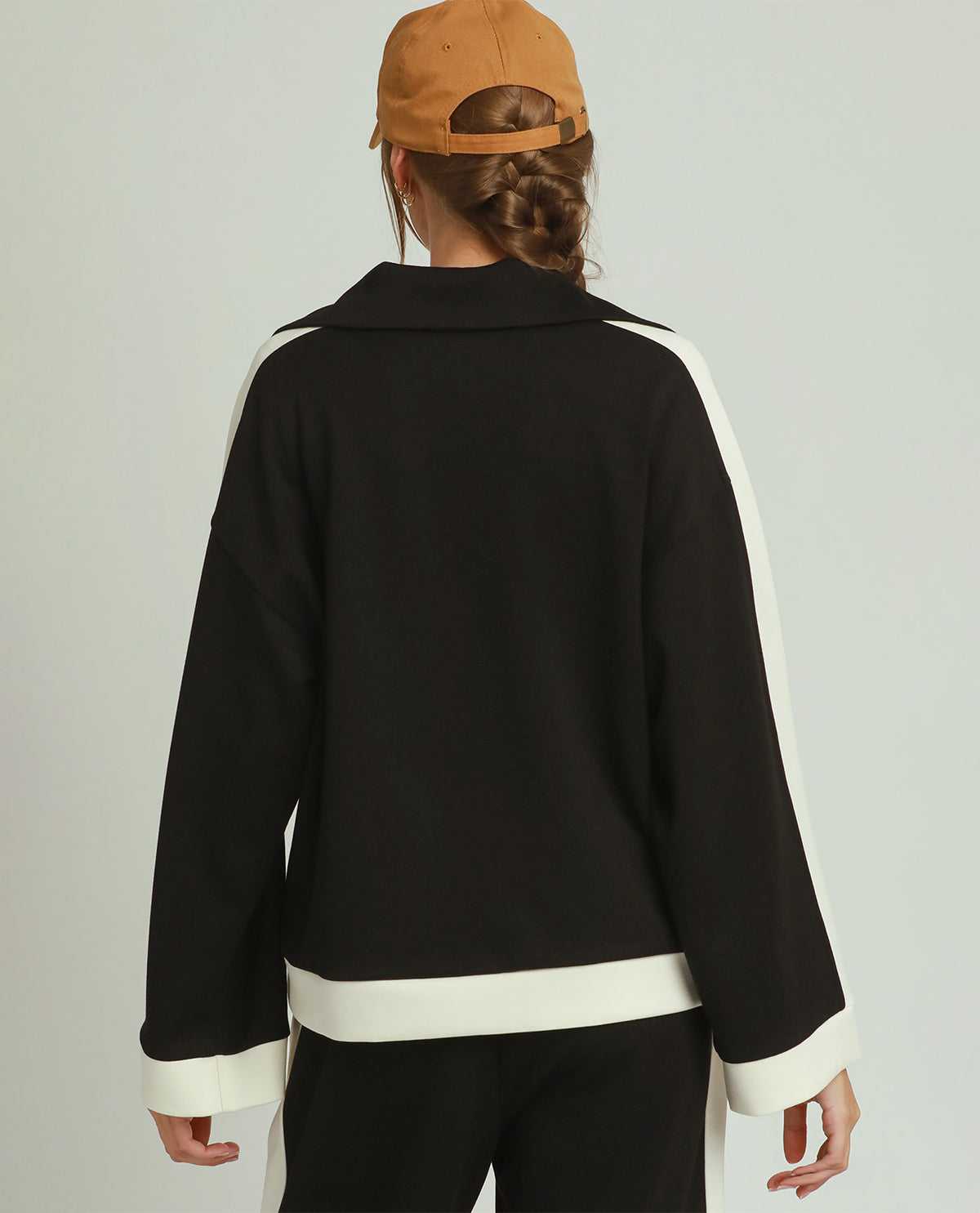 A person shown from the back wearing a black long-sleeve jacket with contrasting white side panels and white trim at the collar, hem, and cuffs, paired with black pants and a tan baseball cap against a light gray studio background.