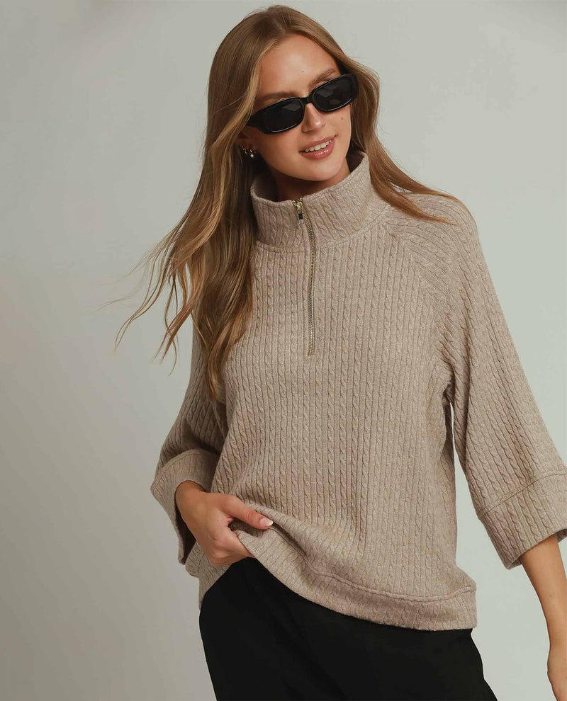 A woman wearing a beige cable-knit mock-neck quarter-zip sweater with three-quarter sleeves, styled with black pants and black sunglasses against a light gray studio background.