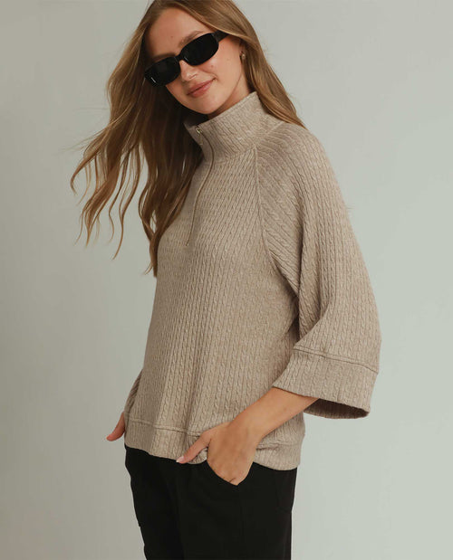 A woman wearing a beige ribbed mock-neck knit sweater with three-quarter sleeves and black pants, posing against a light gray studio background.
