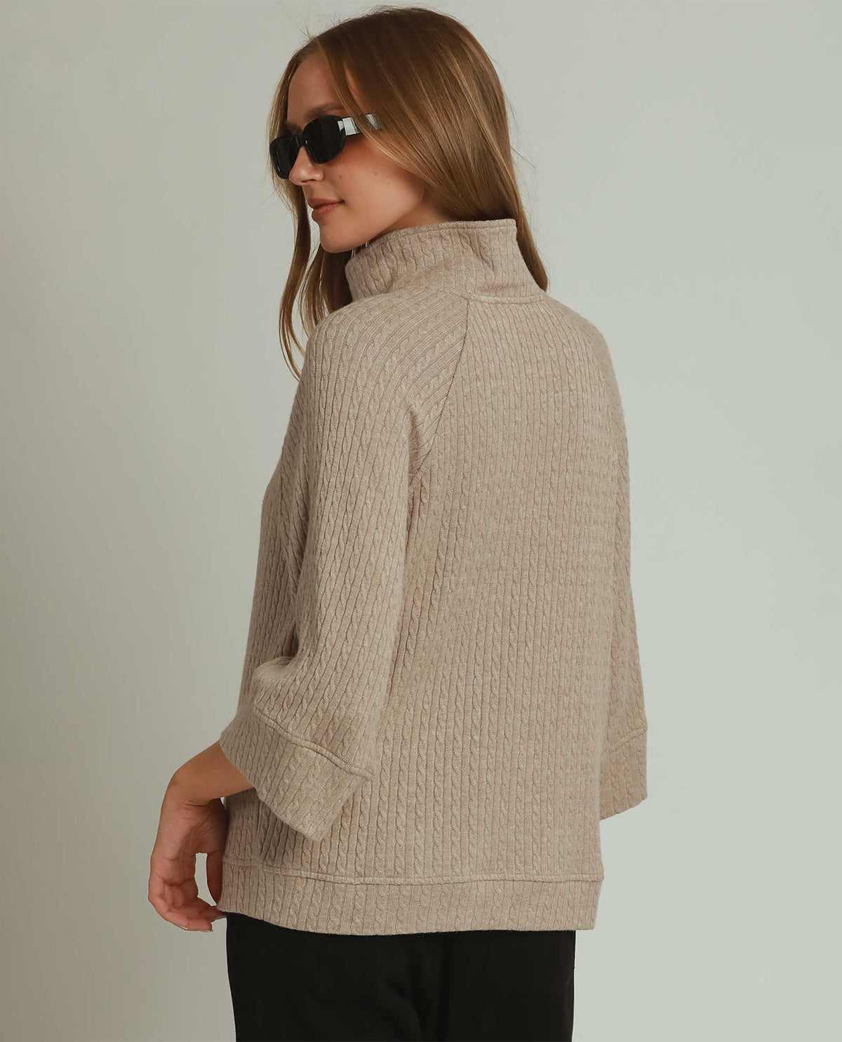 A woman wearing a beige ribbed knit mock neck sweater, shown from a side-back angle against a plain light gray studio background.