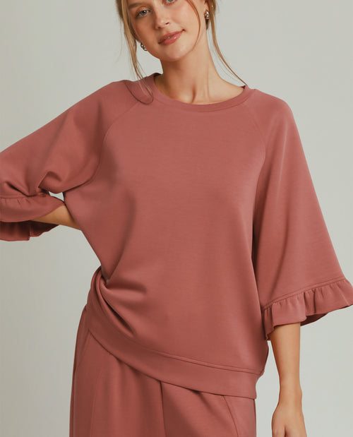A woman wearing a dusty rose pink lounge set featuring a short-sleeve top with ruffled cuffs and matching pants, photographed against a solid light gray background.