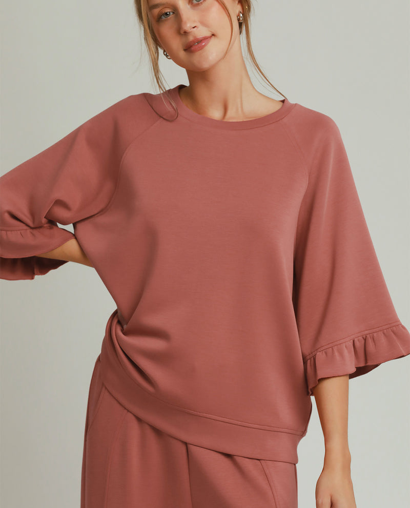 A woman wearing a dusty rose pink lounge set featuring a short-sleeve top with ruffled cuffs and matching pants, photographed against a solid light gray background.