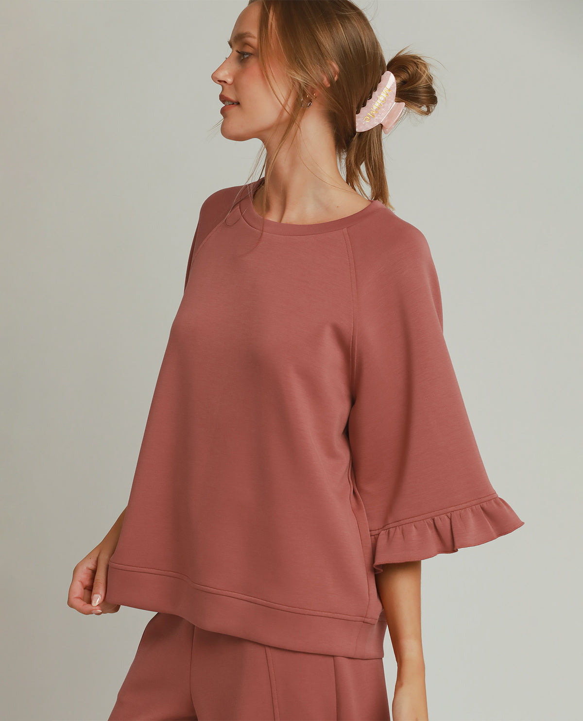 A woman wearing a dusty rose two-piece lounge set, featuring a short-sleeve top with ruffled cuffs and matching pants, photographed against a solid light gray background.