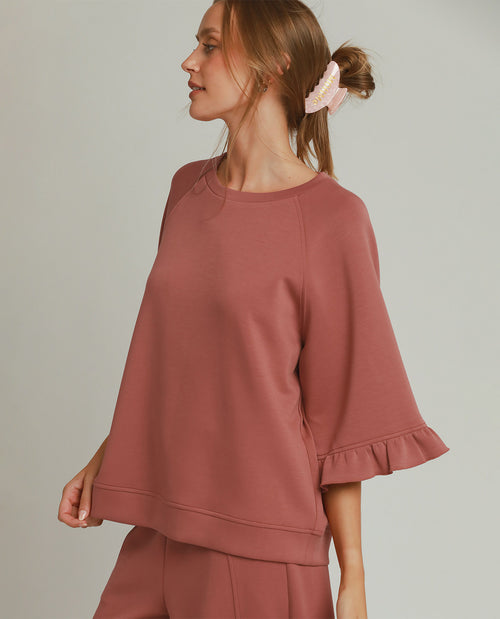 A woman wearing a dusty rose two-piece lounge set, featuring a short-sleeve top with ruffled cuffs and matching pants, photographed against a solid light gray background.