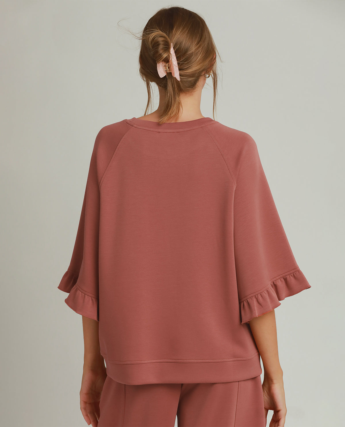A woman shown from the back wearing a dusty rose cropped sweatshirt with ruffled elbow-length sleeves and matching sweatpants against a light gray studio background.