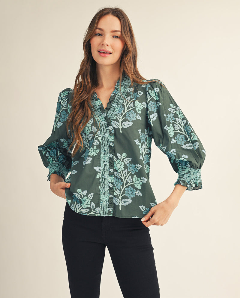 A woman modeling a dark green long-sleeve blouse with a light blue and white botanical pattern, paired with black pants, against a solid beige background.