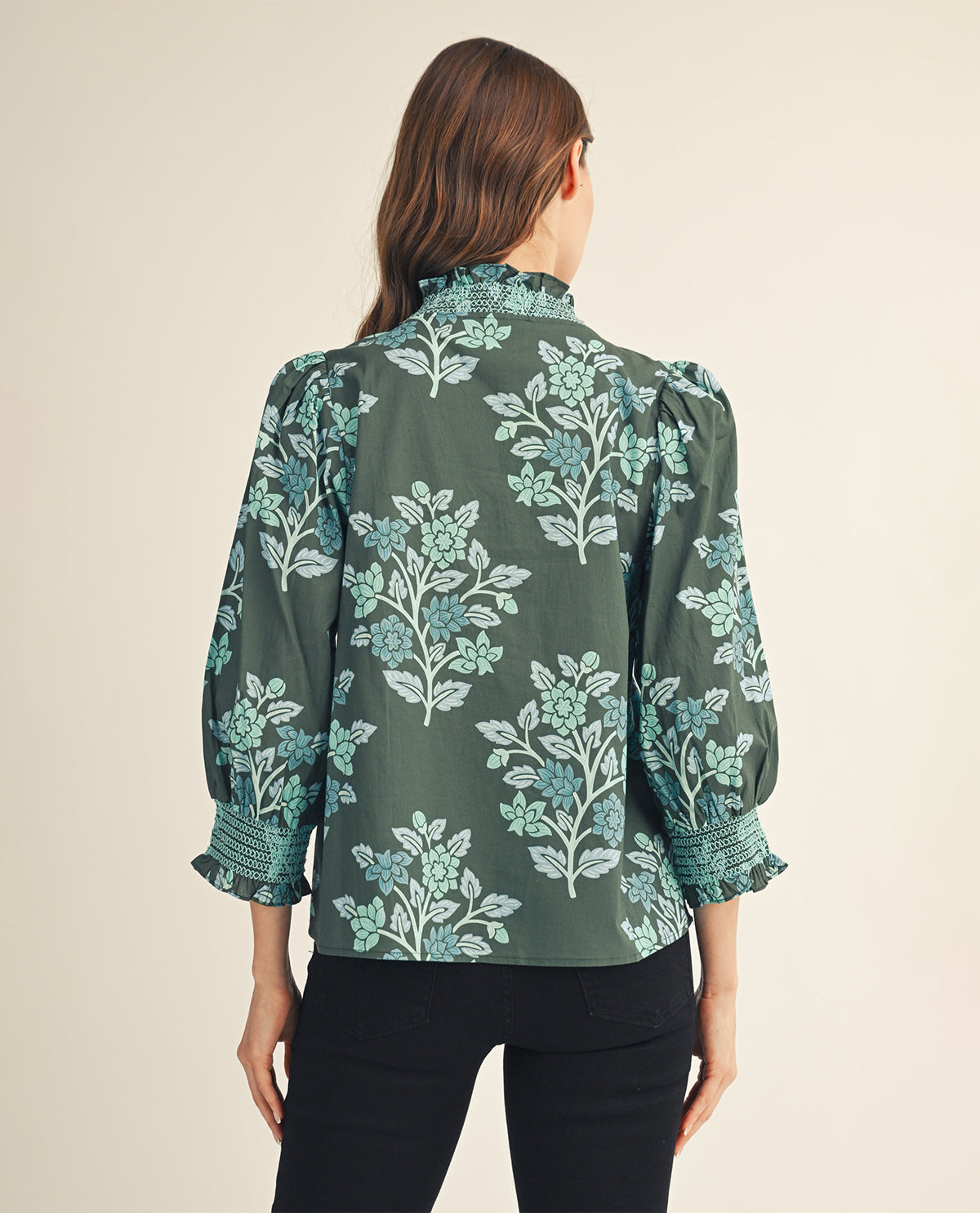 A woman viewed from the back wearing a dark green long-sleeve blouse with a light blue and white floral pattern, paired with black pants, against a solid beige background.