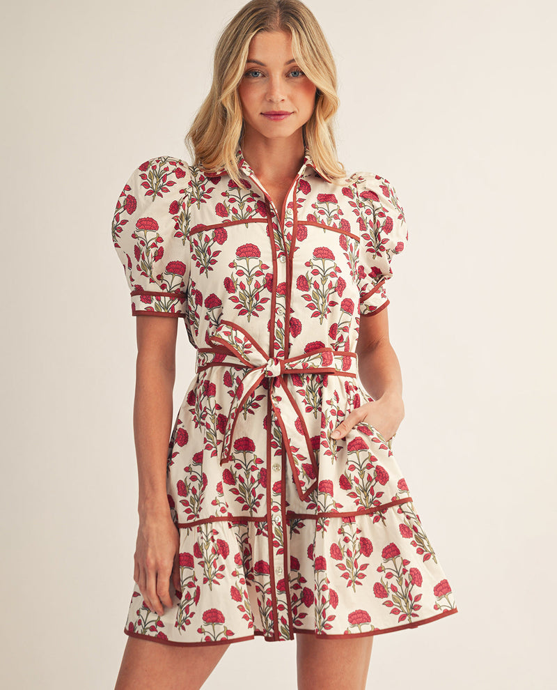 A woman modeling a short puff-sleeve dress with a red and green floral print on a cream background, featuring a matching tie belt at the waist, against a plain light beige studio backdrop.