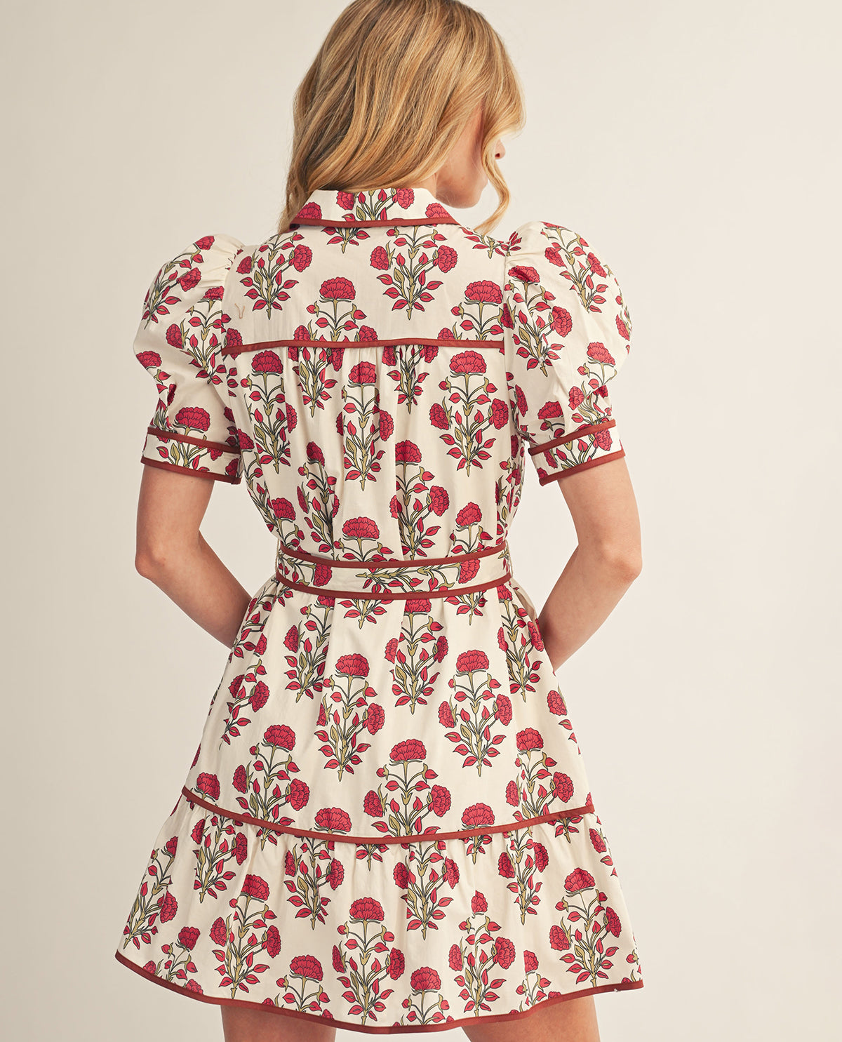 A woman shown from the back wearing a short-sleeve cream dress with a red and green floral print and red trim, against a plain light beige studio background.