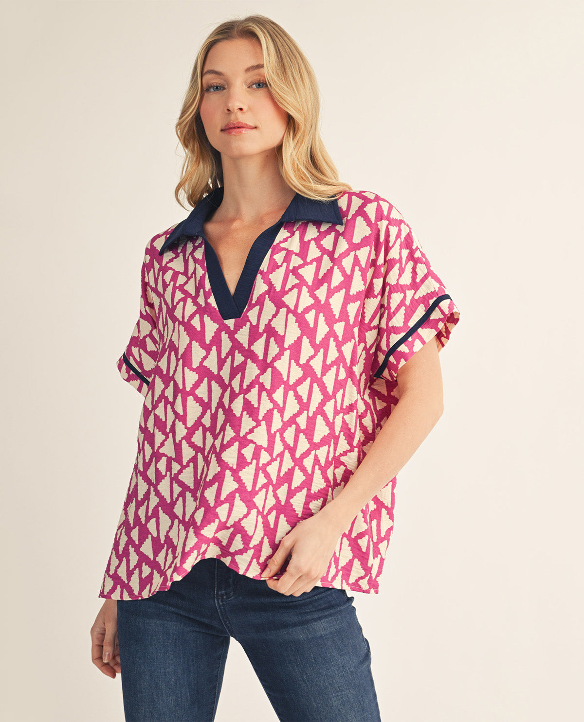 A woman shown from the front wearing a short-sleeve blouse with a pink and cream geometric pattern and dark navy trim, paired with blue jeans against a light beige studio background.