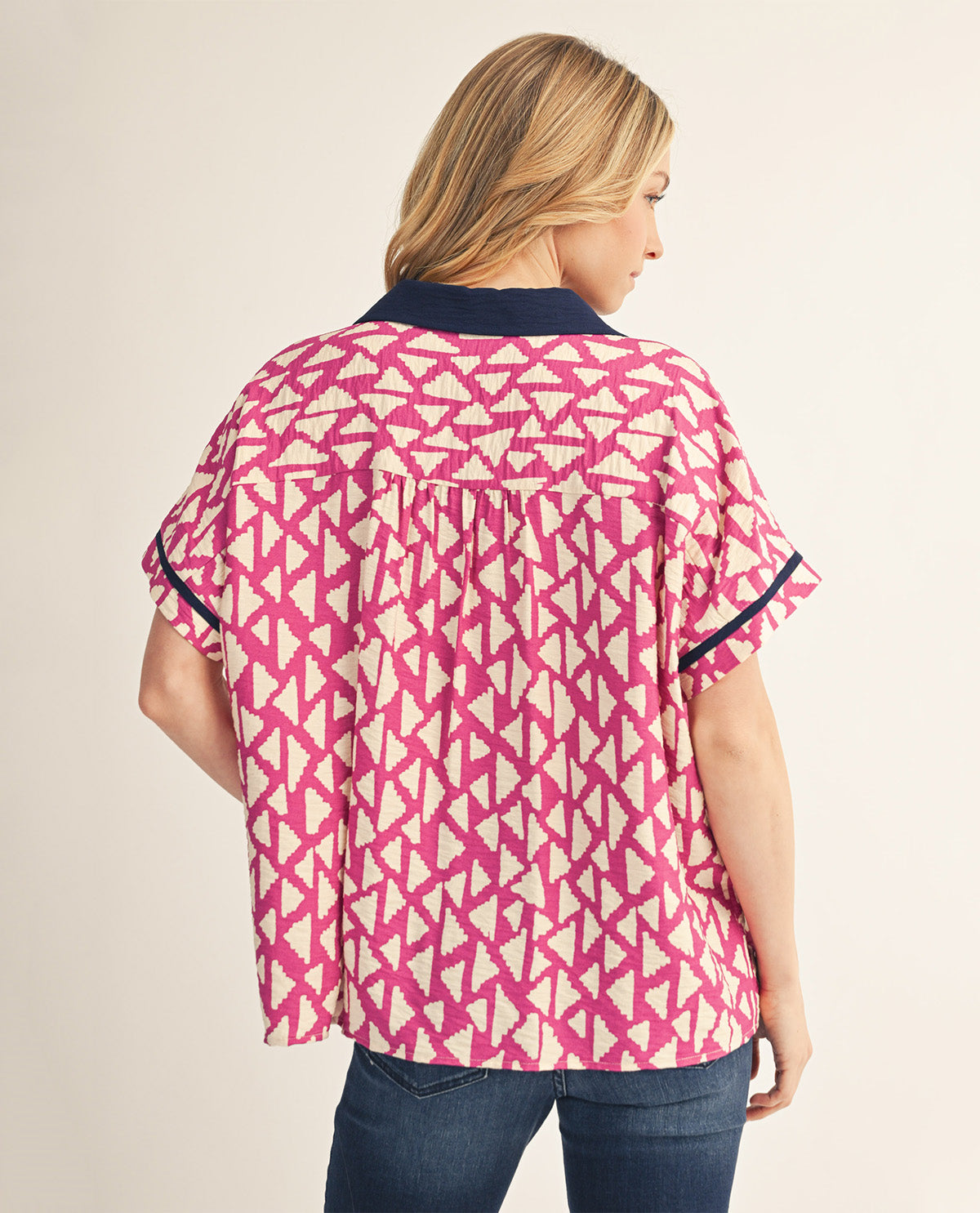 A woman shown from the back wearing a short-sleeve blouse with a pink and cream geometric pattern and dark navy trim, paired with blue jeans against a light beige studio background.