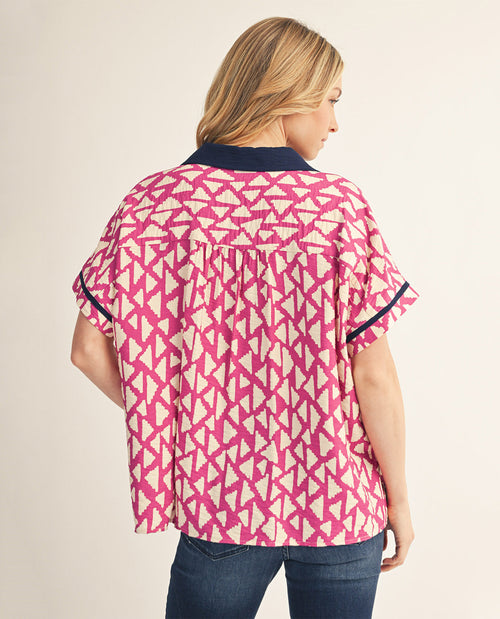 A woman shown from the back wearing a short-sleeve blouse with a pink and cream geometric pattern and dark navy trim, paired with blue jeans against a light beige studio background.