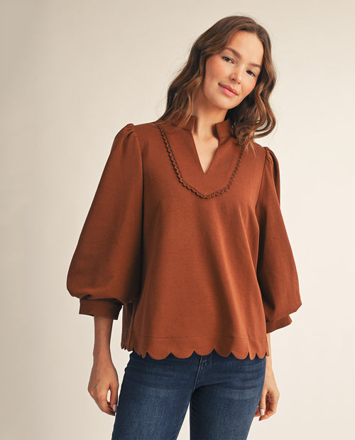 A woman wearing a rust-brown long-sleeve blouse with a scalloped hem and a decorative chain detail along the V-neckline, paired with blue jeans, against a plain beige studio background.