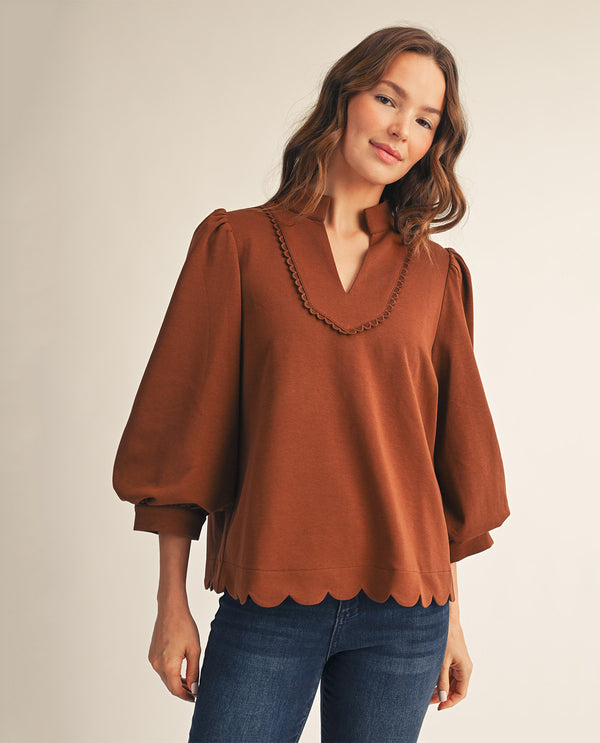 A woman wearing a rust-brown long-sleeve blouse with a scalloped hem and a decorative chain detail along the V-neckline, paired with blue jeans, against a plain beige studio background.