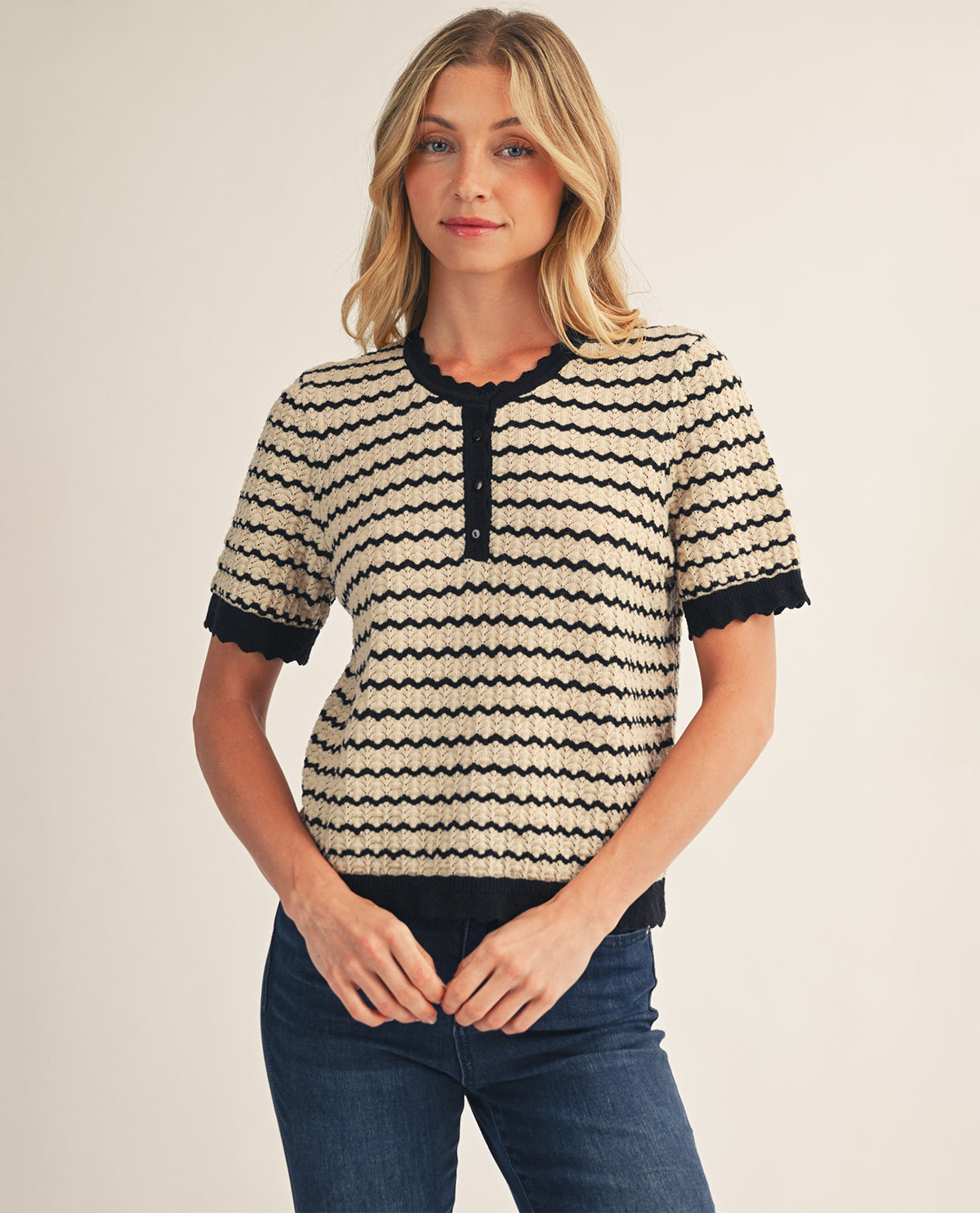 A woman wearing a short-sleeve knit top with a beige and black wavy stripe pattern, featuring a scalloped henley neckline and cuffs, paired with dark blue jeans, against a plain light beige background.