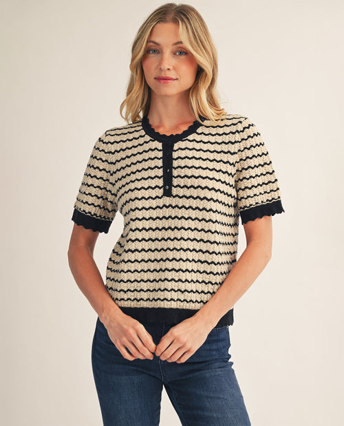 A woman wearing a short-sleeve knit top with a beige and black wavy stripe pattern, featuring a scalloped henley neckline and cuffs, paired with dark blue jeans, against a plain light beige background.