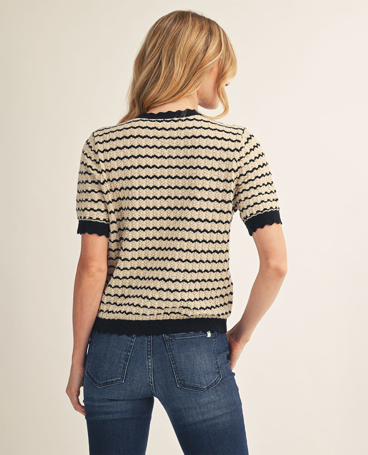 A woman viewed from the back wearing a short-sleeve knit top with horizontal black zigzag stripes on a cream background, paired with blue jeans, against a plain light beige studio background.
