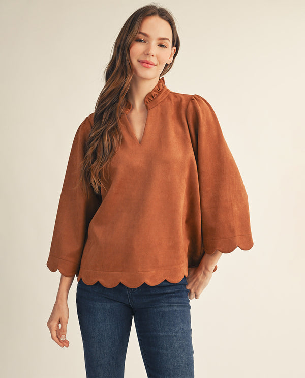 A woman modeling a rust-brown suede-like blouse with a ruffled high neckline, voluminous elbow-length sleeves, and a scalloped hem, paired with dark blue jeans against a plain light beige studio background.