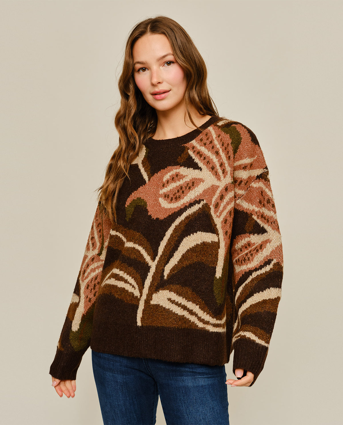 A woman modeling a brown knit sweater with a large abstract floral pattern in beige, pink, and olive green, paired with blue jeans against a solid light gray background.