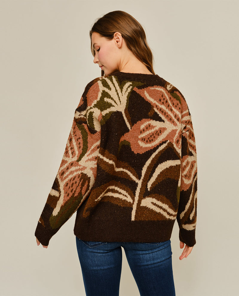 A woman shown from the back wearing a dark brown knit sweater with a large botanical leaf pattern in beige, rust, and brown, paired with blue jeans against a plain light gray studio background.