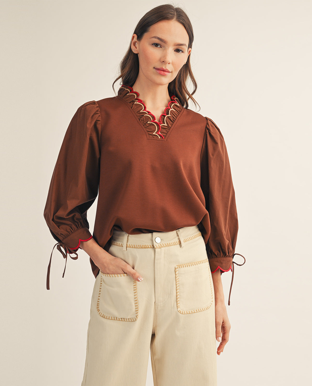 A woman modeling a brown long-sleeve blouse with a decorative red and gold embroidered V-neckline and tie details at the cuffs, paired with cream-colored pants, against a plain light beige background.