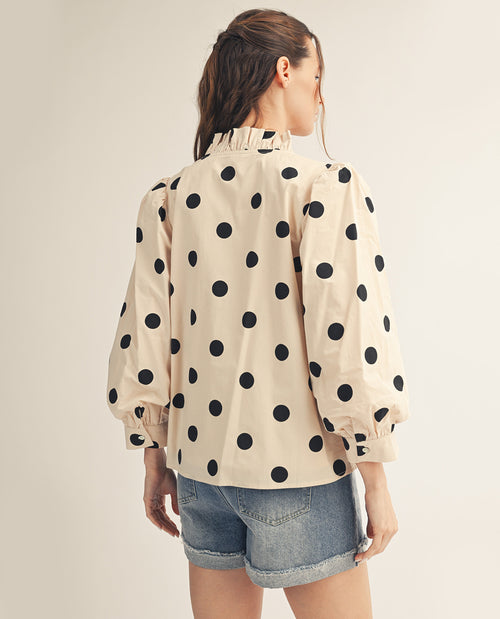 Woman wearing a beige blouse with black polka dots and denim shorts on a plain background