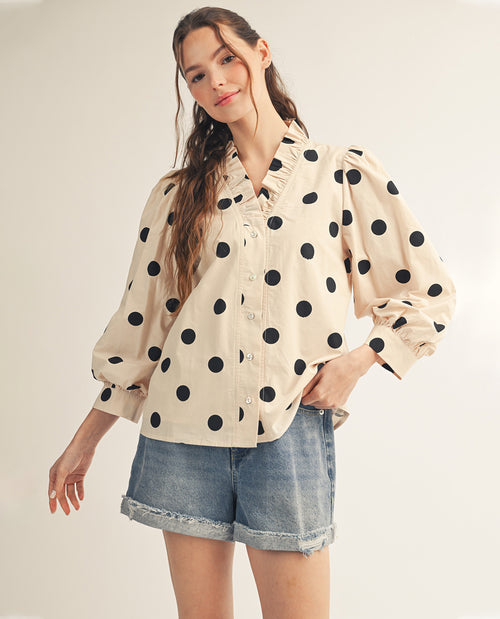 Woman wearing a beige blouse with black polka dots and denim shorts on a plain background