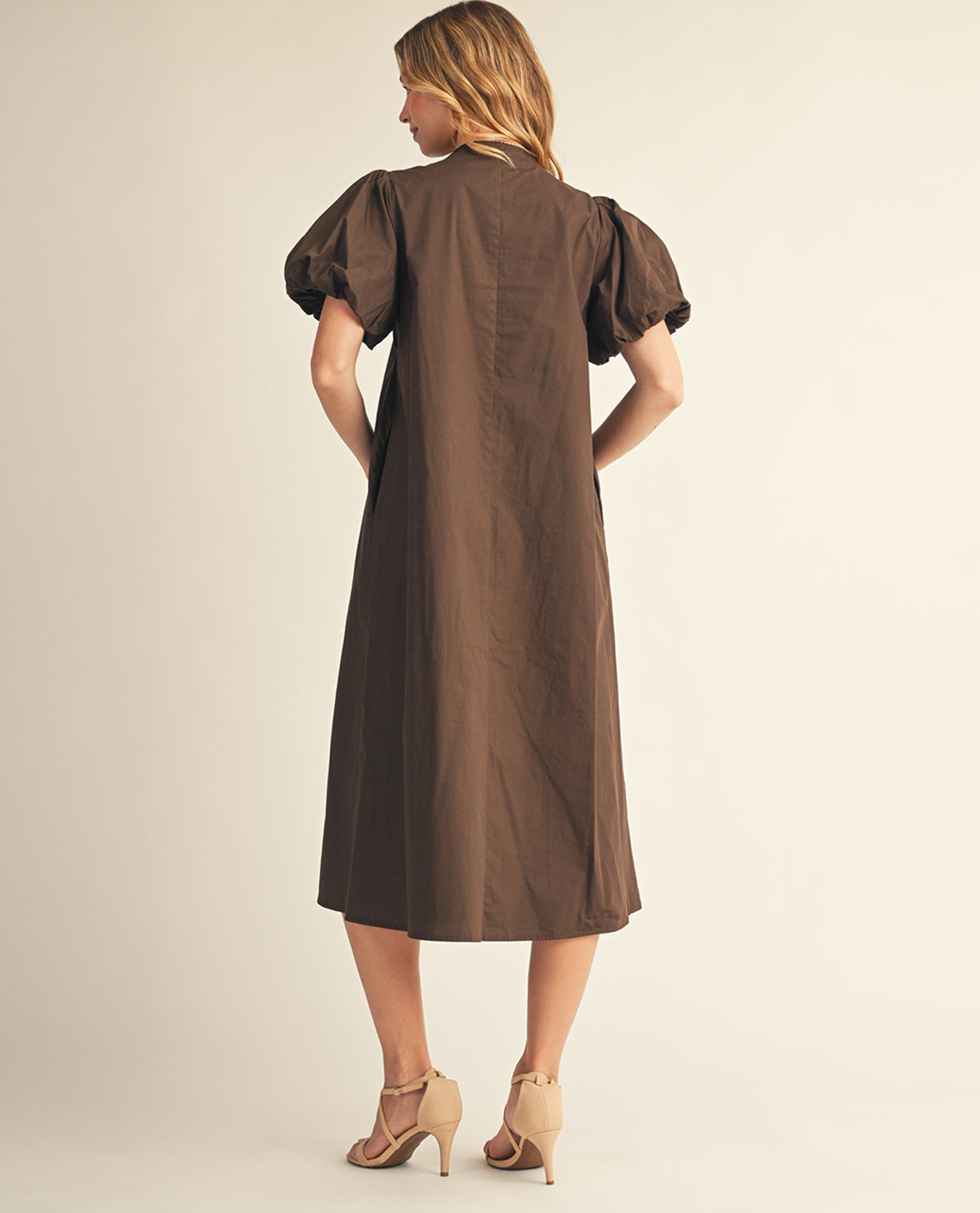 A woman shown from the back wearing a brown midi dress with puffed short sleeves and beige high-heeled sandals, standing against a plain light beige studio background.